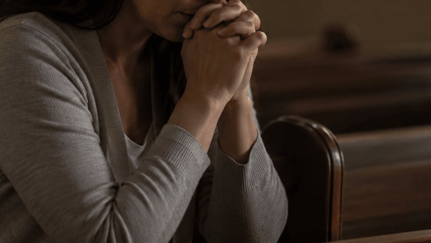 Mulher de mãos unidas junto ao rosto, sentada em um banco de igreja.