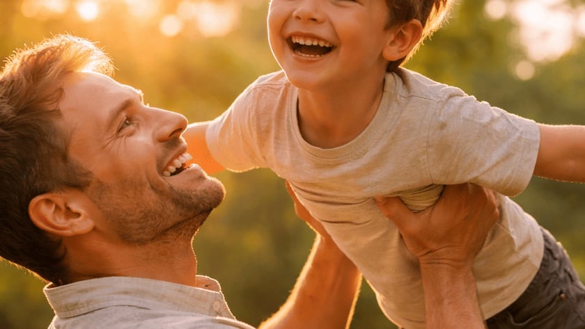 Homem sorridente levanta um menino com os braços abertos, ao ar livre.