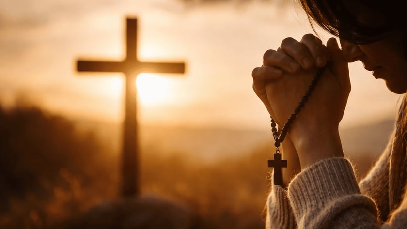 Mulher de suéter claro, de cabeça baixa, reza com um terço nas mãos diante de uma cruz.