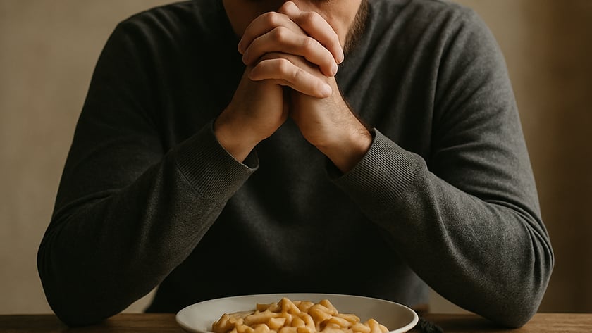 Homem de blusa cinza reza com as mãos unidas diante de um prato de macarrão.