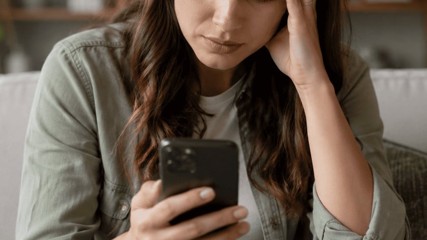 Mulher de cabelo escuro sentada no sofá, olhando o celular com a mão na testa.