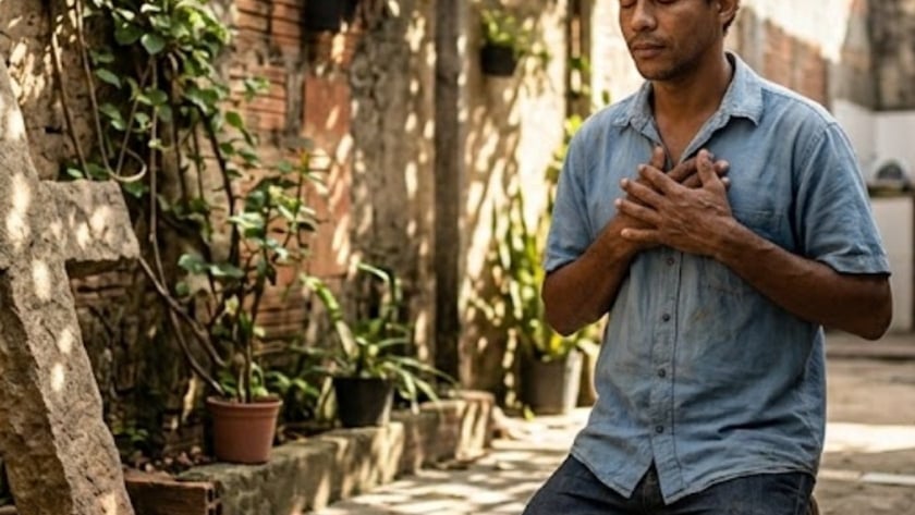 Homem ajoelhado com as mãos no peito e olhos fechados, ao lado de uma cruz de pedra, em um quintal.