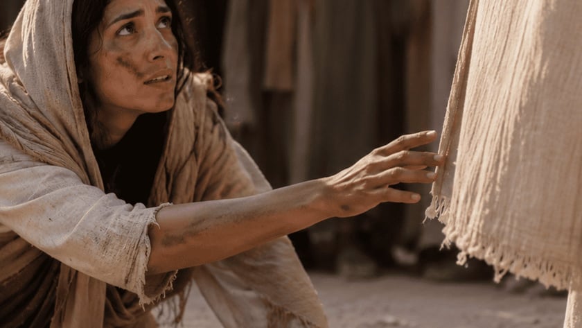 Mulher ajoelhada na terra estende a mão para tocar a barra de uma túnica clara.