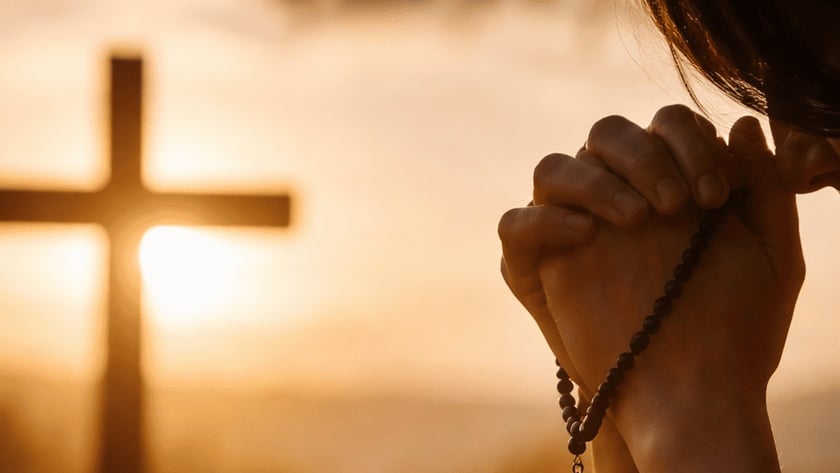 Mulher de suéter claro, de cabeça baixa, reza com um terço nas mãos diante de uma cruz.