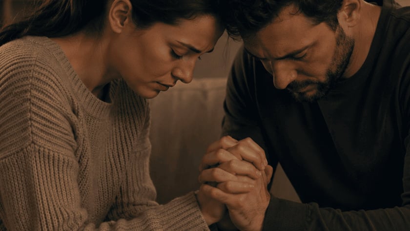 Homem e mulher sentados no sofá, de cabeça baixa, com as mãos unidas.