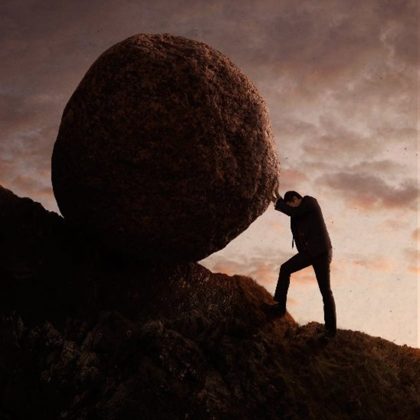 Homem de terno empurra uma rocha gigantesca morro acima ao entardecer.