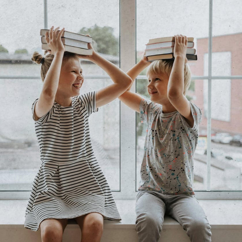 Duas crianças sentadas no parapeito de uma janela, cada uma com uma pilha de livros na cabeça.