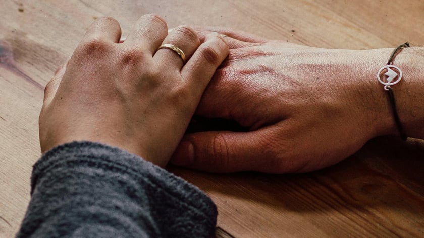 Duas mãos sobrepostas em uma mesa de madeira, uma com aliança dourada.