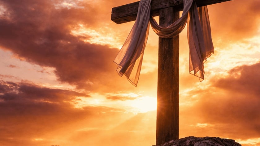 Cruz de madeira com tecido claro pendurado, sobre uma rocha ao pôr do sol.