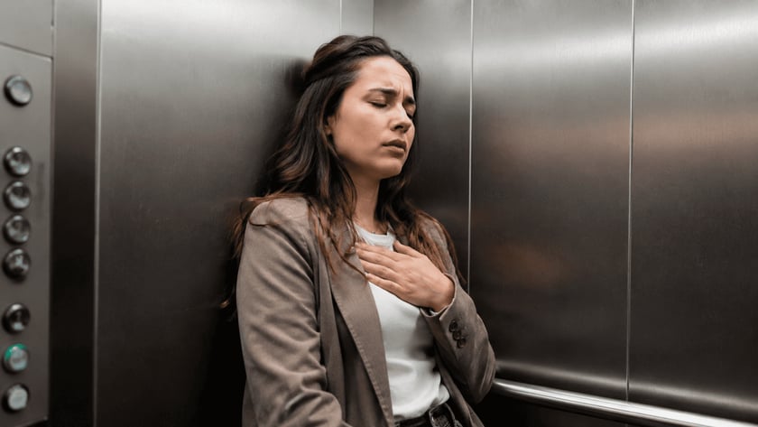 Mulher de blazer marrom encostada na parede de um elevador, com a mão no peito.