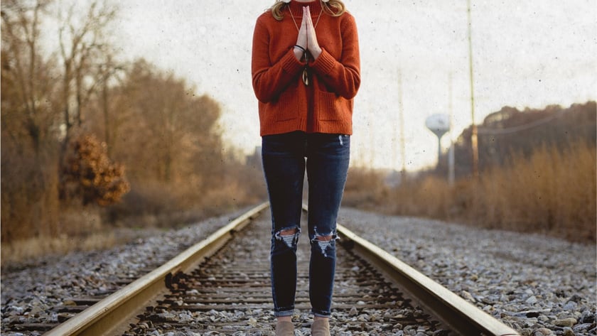 Mulher de suéter laranja e jeans rasgado, com as mãos juntas, em pé sobre trilhos de trem.