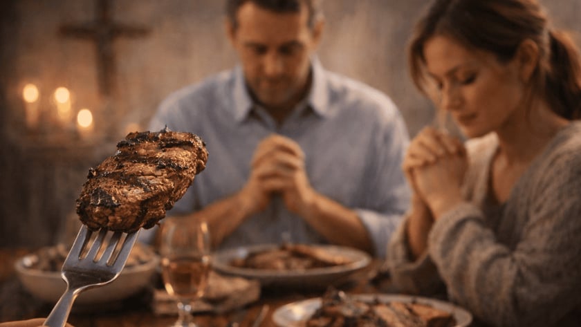 Homem de mãos postas, de cabeça baixa, à mesa com pratos de carne e um garfo em primeiro plano.