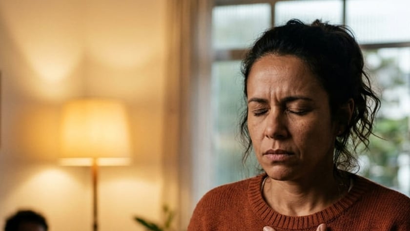 Mulher de suéter marrom com a mão no peito e os olhos fechados, em uma sala.