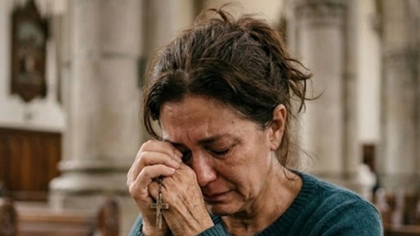 Mulher de suéter azul segura um terço junto ao rosto, sentada num banco de igreja.