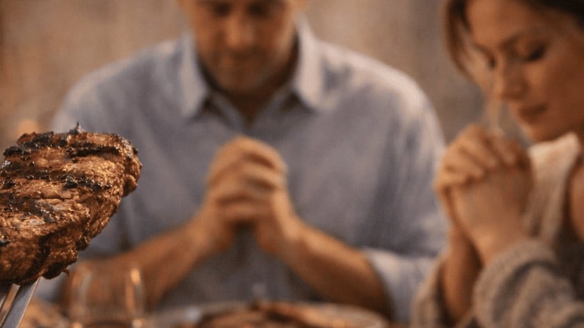 Homem de mãos postas, de cabeça baixa, à mesa com pratos de carne e um garfo em primeiro plano.