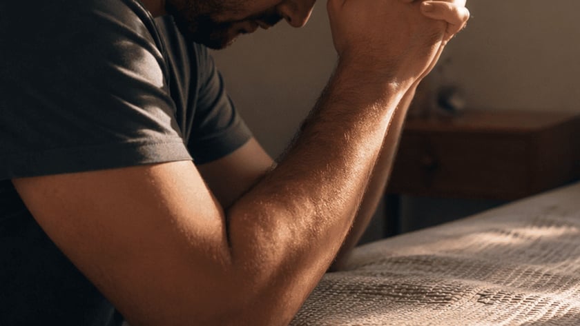 Homem de camiseta cinza, de olhos fechados e mãos postas na testa, ao lado de uma cama.