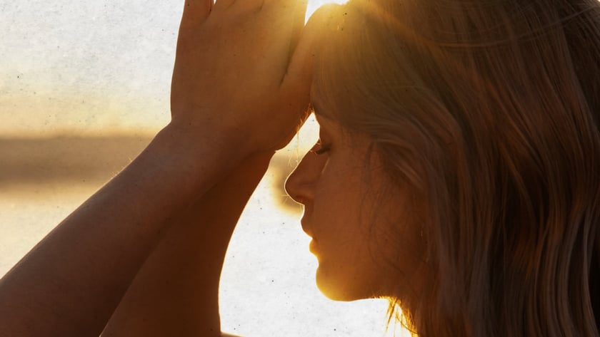 Mulher de perfil, com cabelo curto, une as palmas das mãos acima da testa ao pôr do sol.