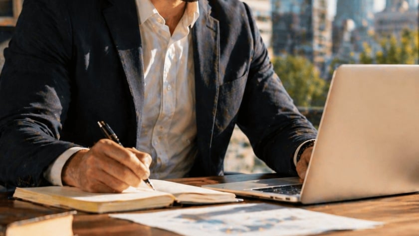Homem de blazer escreve em caderno enquanto usa notebook, com prédios ao fundo.