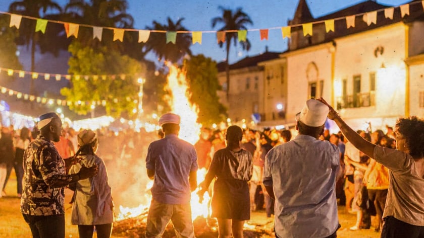 Pessoas de costas dançam ao redor de uma fogueira numa praça com bandeirinhas coloridas.