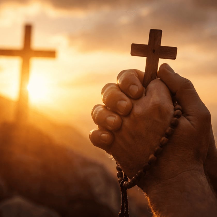 Mãos postas segurando um terço com crucifixo de madeira diante de uma cruz ao pôr do sol.