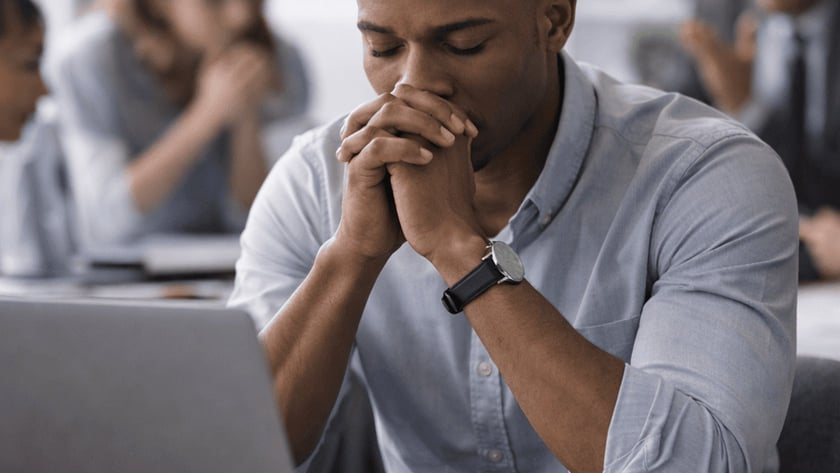Homem de camisa azul com as mãos unidas diante do rosto, sentado diante de um notebook.