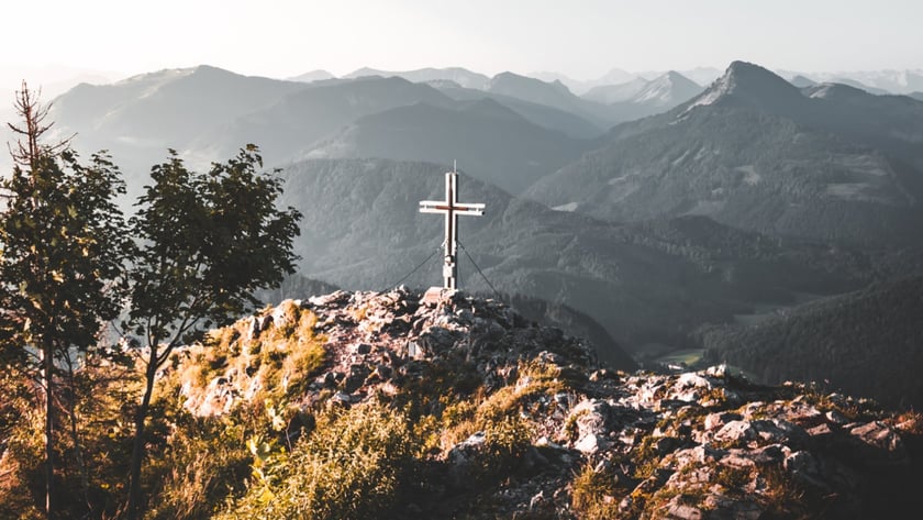 Cruz de metal no topo rochoso de uma montanha, com cadeias de montanhas ao fundo.