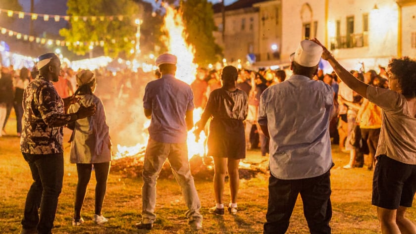 Pessoas de costas dançam ao redor de uma fogueira numa praça com bandeirinhas coloridas.