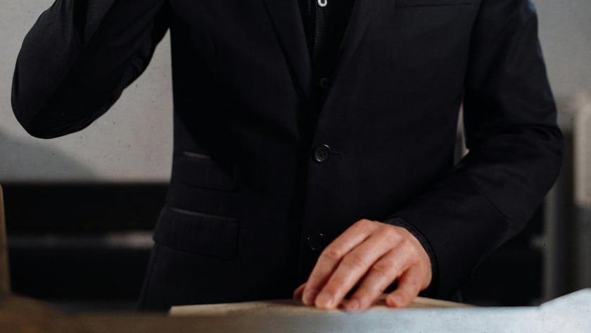Homem barbudo de terno preto ergue os óculos e olha papéis sobre uma mesa.