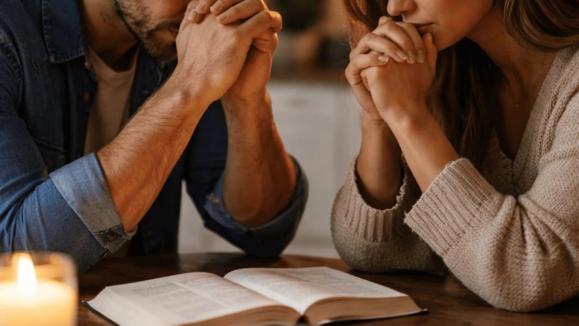 Homem e mulher de mãos unidas e olhos fechados rezando à mesa, com Bíblia aberta.