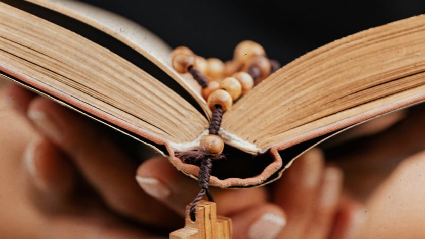 Mãos seguram um livro aberto com um terço de contas de madeira e cruz.